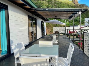 Une terrasse extérieure avec une table à plateau de verre et des chaises blanches, une pergola avec un toit de chaume et une clôture blanche agrémentée de panneaux décoratifs en treillis.