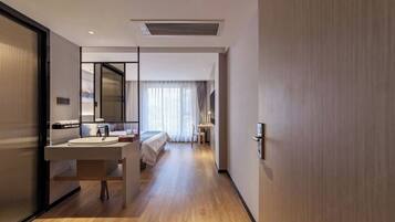 A modern hotel room with a wooden floor, a large mirror, a bed, a bedside table, and a bathroom with a sink and mirror.