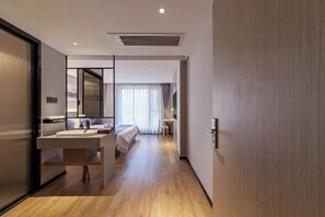 A modern hotel room with a wooden floor, a large mirror, a bed, a bedside table, and a bathroom with a sink and mirror. - Allston Hotel (Xiamen)
