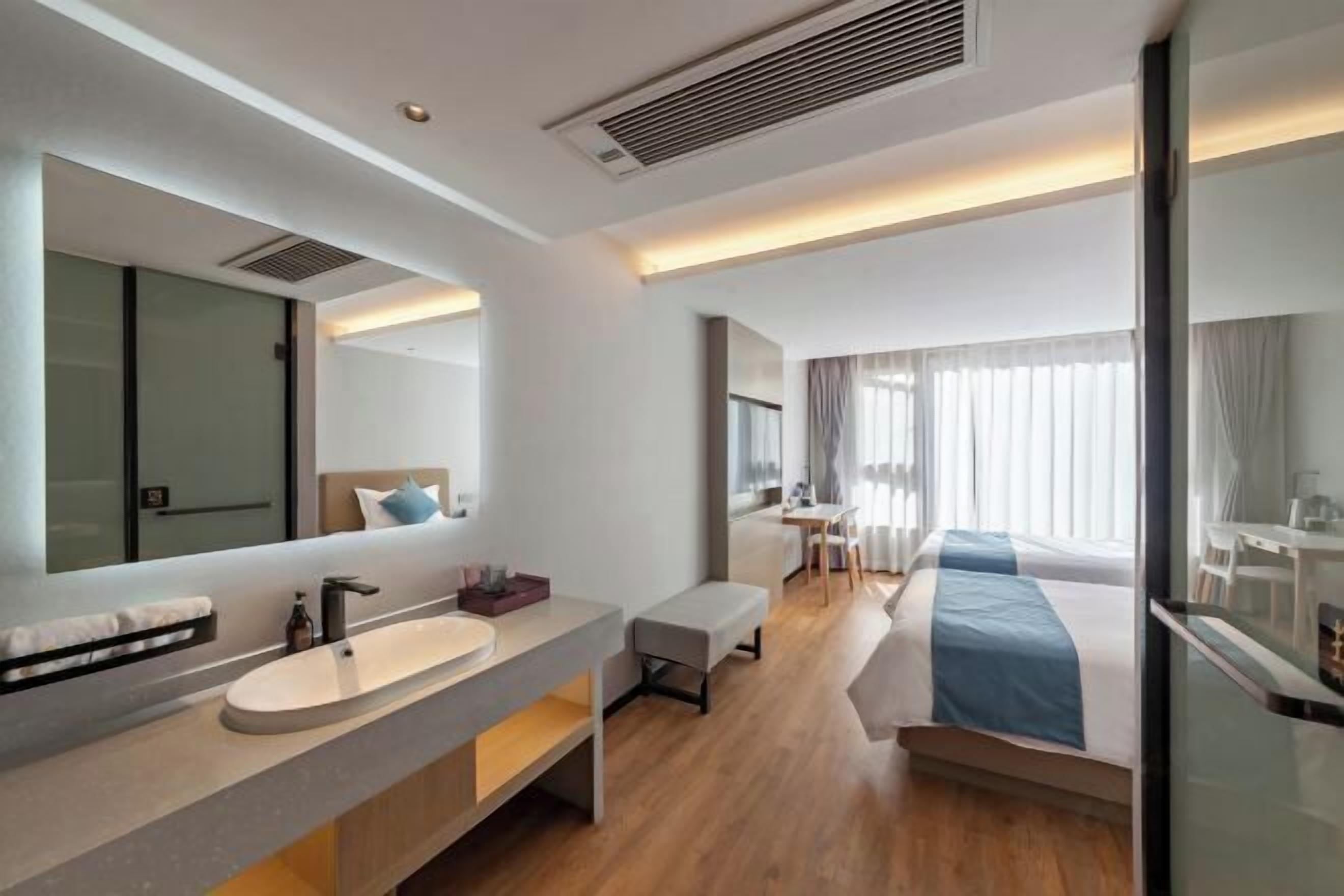 A modern hotel room with a large mirror, a round sink, a bedside table, a bed with blue bedding, a small desk, and a chair.