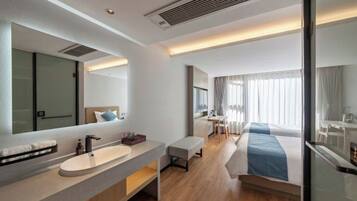 A modern hotel room with a large mirror, a round sink, a bedside table, a bed with blue bedding, a small desk, and a chair.