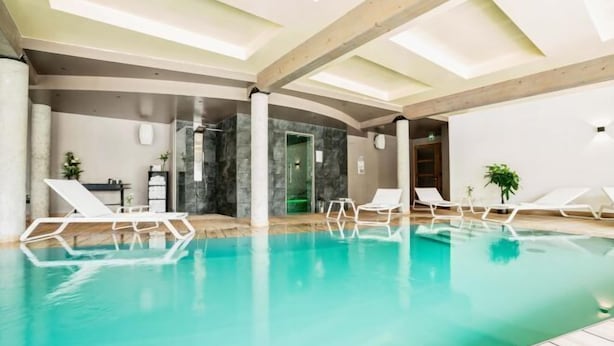 An indoor swimming pool with clear water, white lounge chairs, and a modern architectural design with exposed beams and stone walls.