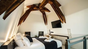 A modern bedroom with a sloped ceiling, a bed with white and dark bedding, a desk with a computer, and a TV mounted on the wall.