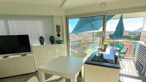 En moderne stue med et fladskærms-TV, hvide møbler, en balkon med udsigt over byen og et stort vindue med en blå parasol.
