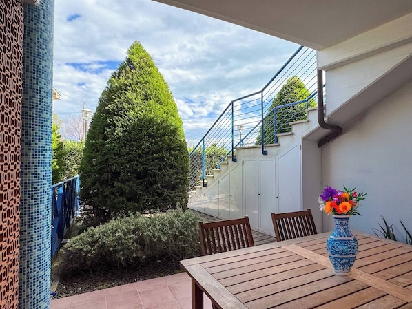 A patio with a wooden table and chairs, a blue vase with flowers, and a tall coniferous tree.