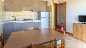 A compact kitchen with a gas stove, sink, and refrigerator. There is a wooden dining table with chairs. A television is on a wooden cabinet to the right.
