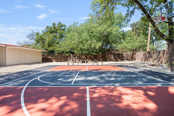 Un terrain de basket en plein air, entouré d’une clôture en bois, avec des arbres et un bâtiment en arrière-plan.