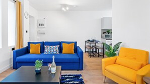 A modern living room with a blue sofa, an orange armchair, a coffee table with a bottle and plant, and a dining area with a table and chairs. - 5 Guests - 2 Bedrooms - Brighton Centre - Petsok (Brighton)