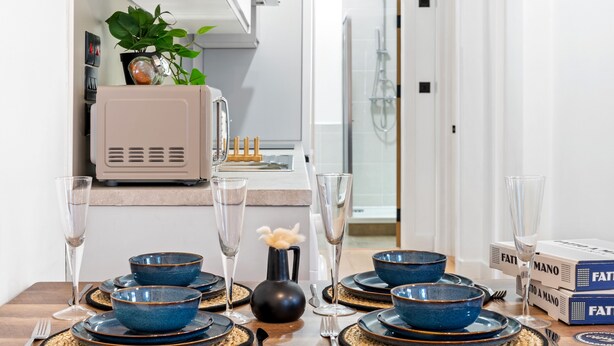 A dining table set with blue bowls, plates, and wine glasses, with a microwave and a plant in the background. - 5 Guests - 2 Bedrooms - Brighton Centre - Petsok (Brighton)