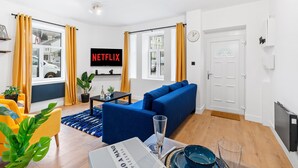 A modern living room with a blue sofa, yellow curtains, a flat-screen TV, and a dining area with a newspaper and glassware. - 5 Guests - 2 Bedrooms - Brighton Centre - Petsok (Brighton)