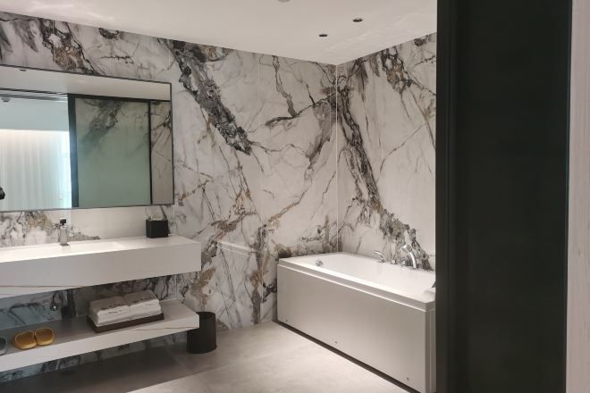 A modern bathroom with a marble wall, a large mirror, a bathtub, and a shelf with toiletries.