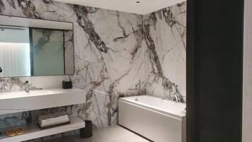 A modern bathroom with a marble wall, a large mirror, a bathtub, and a shelf with toiletries.