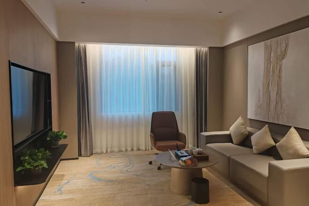 Moderni hotellihuone, jossa on taulutelevisio, sohva, sohvapöytä, tuoli ja suuri ikkuna, jossa on verhot.