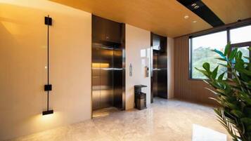 A modern elevator with a metallic finish, a marble floor, and a large window providing a view of greenery outside.