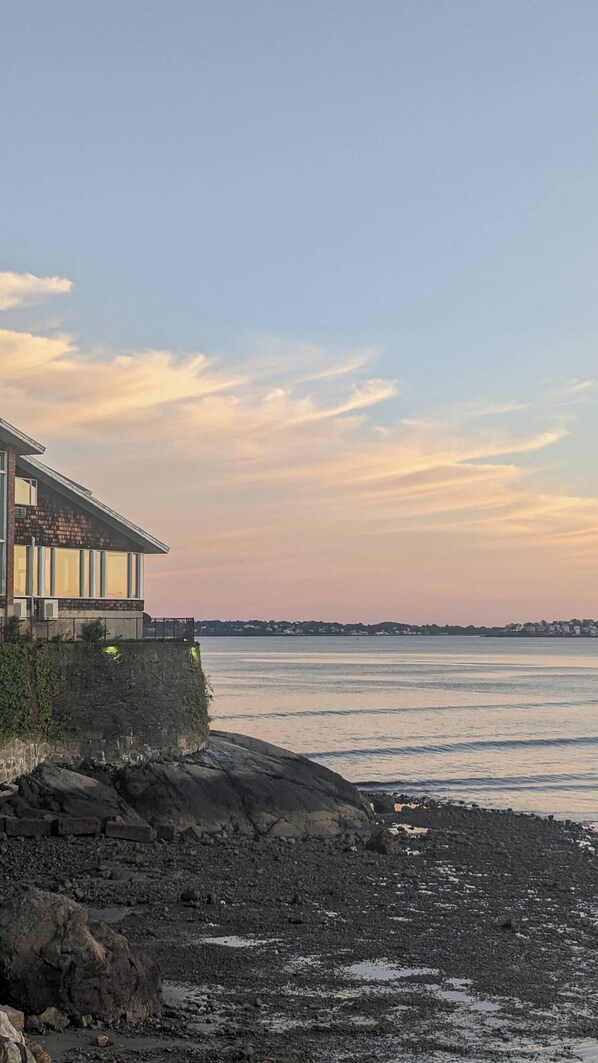A coastal building with a balcony overlooking a rocky shoreline and calm sea.