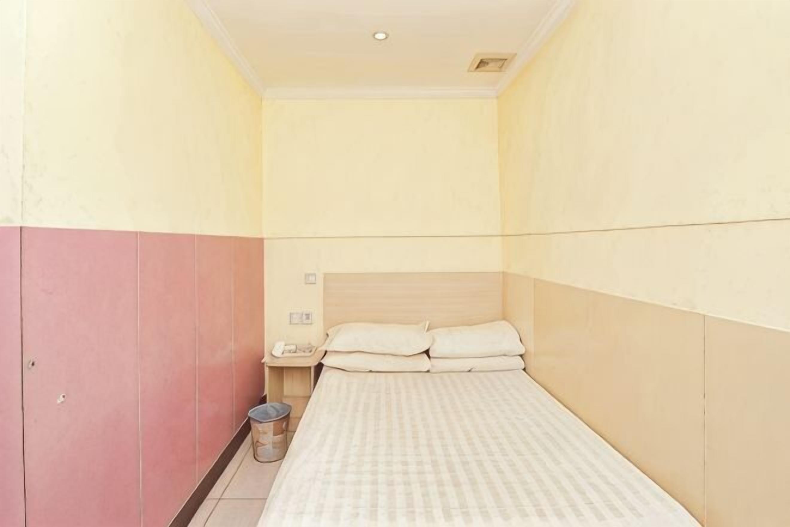 Una habitación pequeña con cama individual, cabecera de madera, mesita de noche y un cubo de basura.