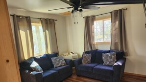 A room with two dark blue sofas, patterned throw pillows, a wooden floor, a ceiling fan, and a window with curtains. - Mountain Cabin in Palmer, Ak (Palmer)