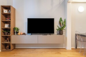 A modern living room with a flat-screen TV mounted on a white wall, wooden shelving unit with decorative items, and a potted plant.