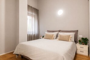 A bedroom with a bed, bedside tables, a nightstand, a window with curtains, and a wall-mounted light.