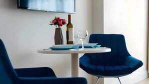 Ein modernes Zimmer mit einem an der Wand befestigten Flachbildfernseher, einem runden Tisch mit Blumen in einer Vase und einer Weinflasche, sowie zwei blauen Stühlen.