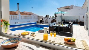 A poolside dining area with a table set for two, a view of the pool, and a white building with a tiled roof in the background. - R1385 | Villa Emma, La Exótica (Nerja)