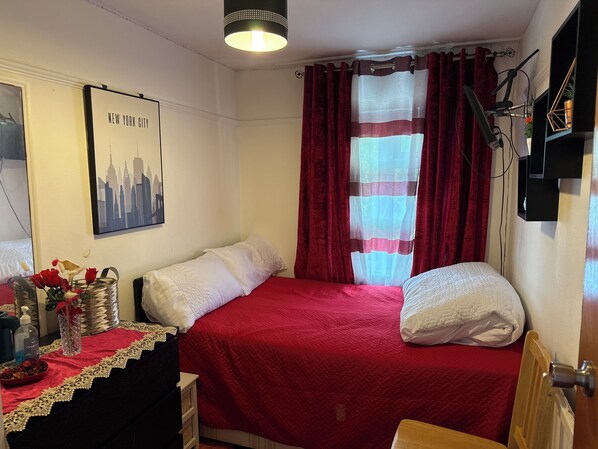 Una habitación con dos camas, una colcha roja y blanca, un cuadro enmarcado con una vista urbana, una ventana con cortinas rojas y una mesita de noche con un jarrón de flores.