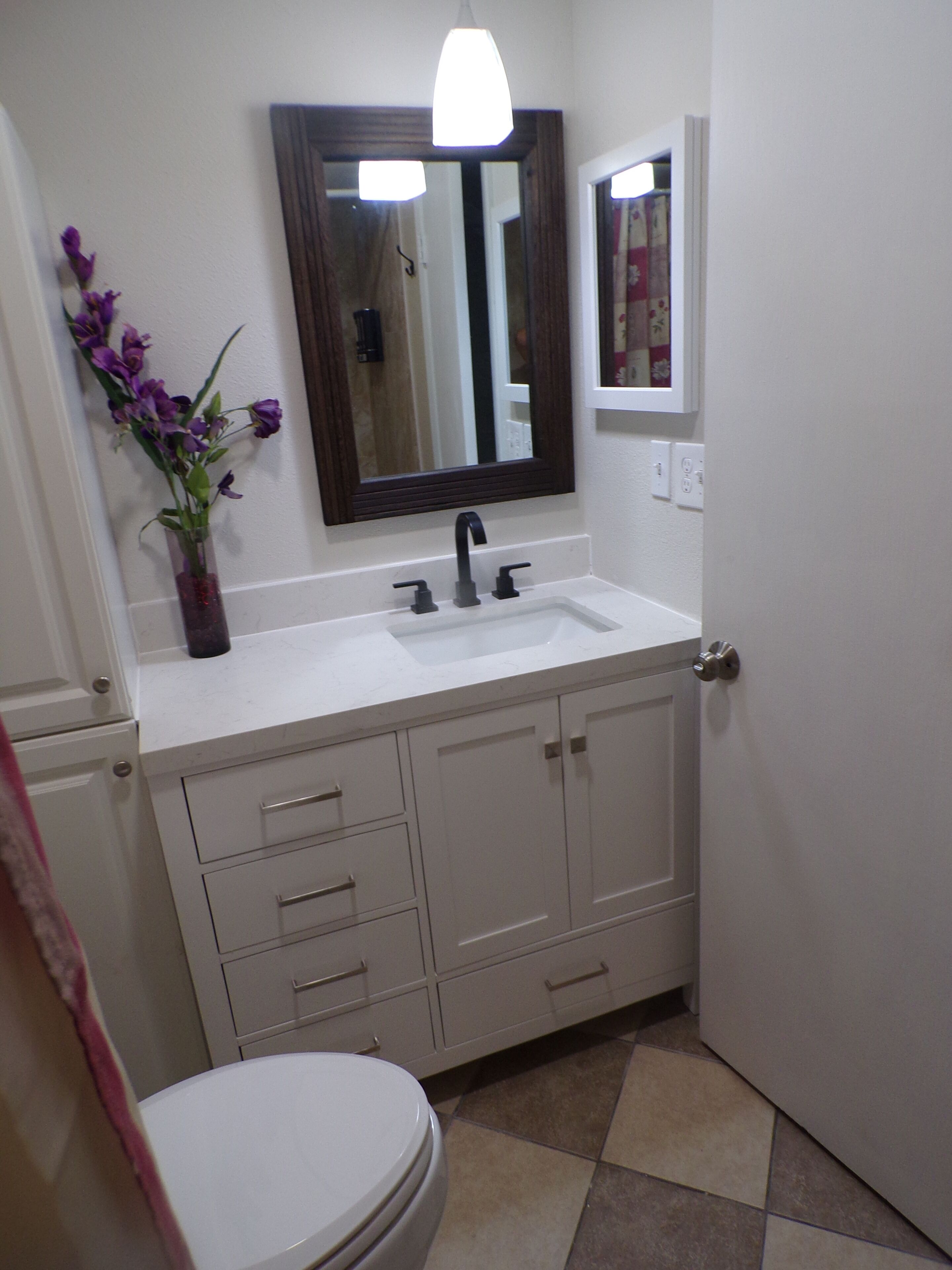 Une salle de bain avec un lavabo blanc, un miroir au-dessus, et des toilettes. Il y a des placards sous le lavabo et un vase avec des fleurs violettes sur la gauche.