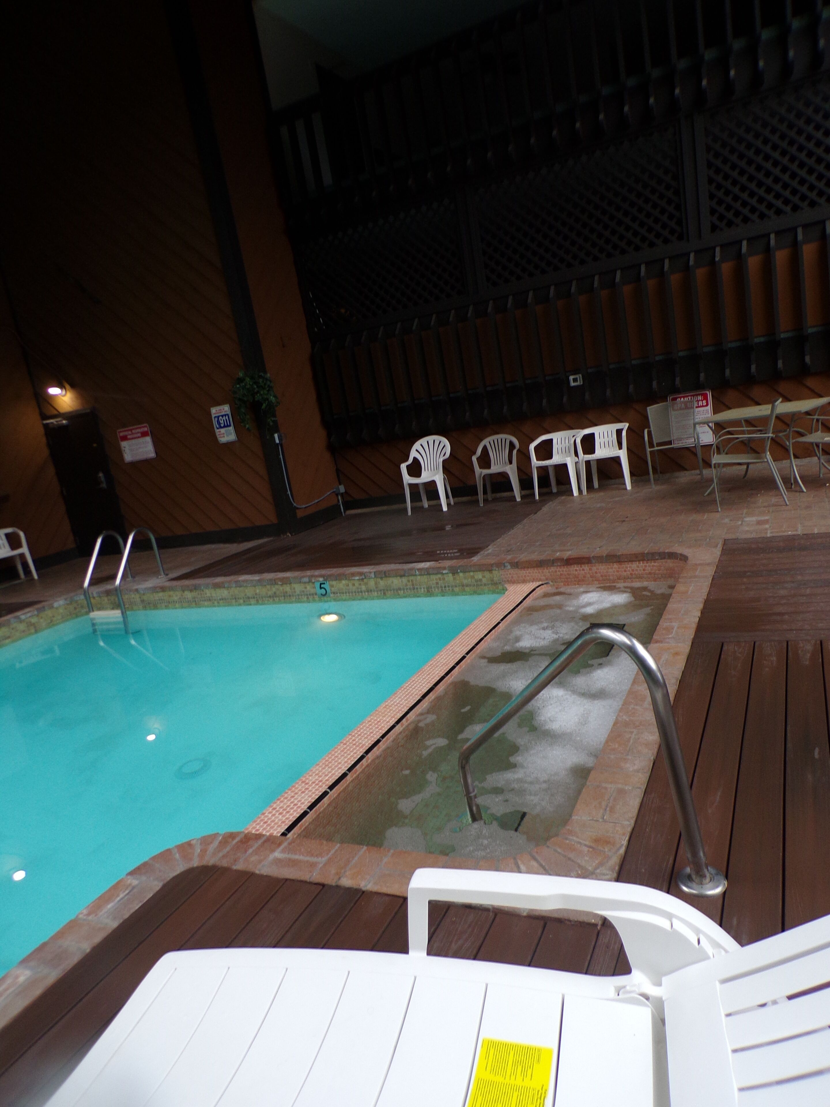 An indoor swimming pool with a wooden deck, white plastic chairs, and a metal ladder.