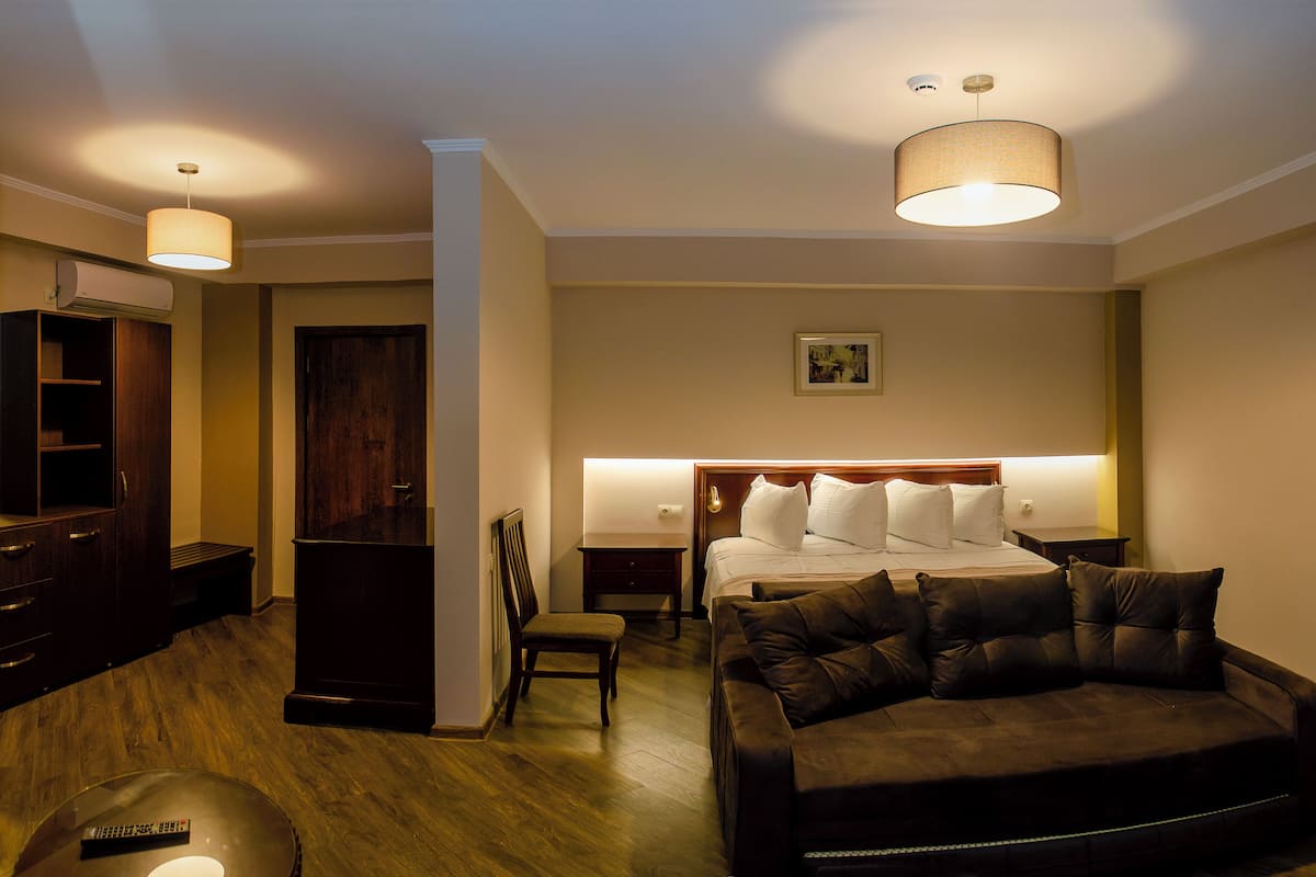 Habitación de hotel con cama, sofá de cuero marrón oscuro, suelo de madera, armario de madera y lámpara en el techo.