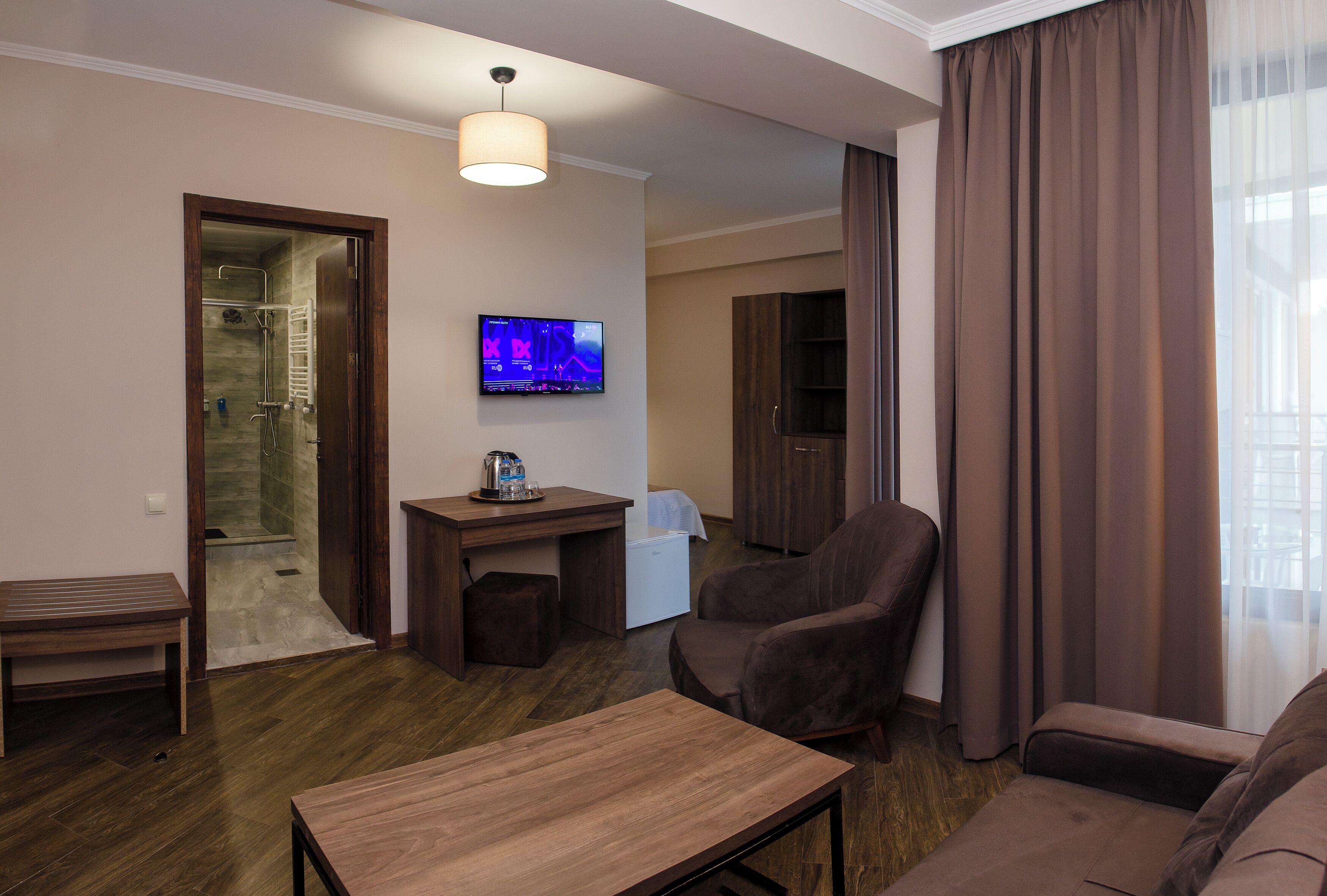 A hotel room with a wooden floor, a TV mounted on the wall, a desk with a lamp, a chair, a sofa, and a shower area visible through an open door.