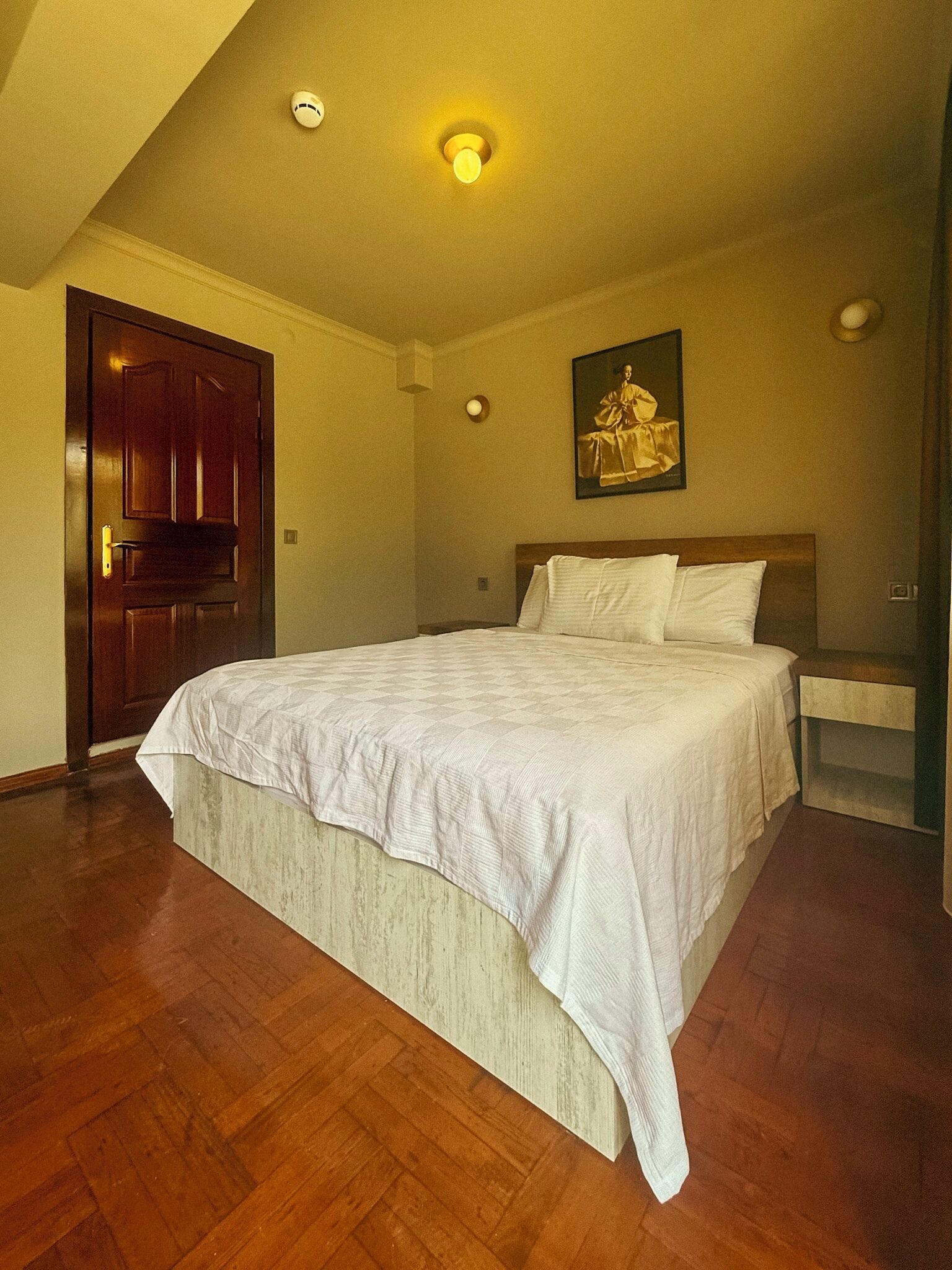 A bedroom with a wooden bed, a nightstand, a painting on the wall, and a door.