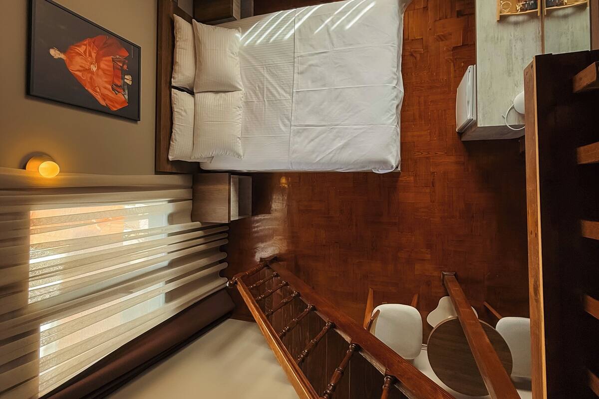Ein Schlafzimmer mit Bett, einem hölzernen Kopfteil, einem Nachttisch, einem gerahmten Bild und einem Fenster mit Jalousien.