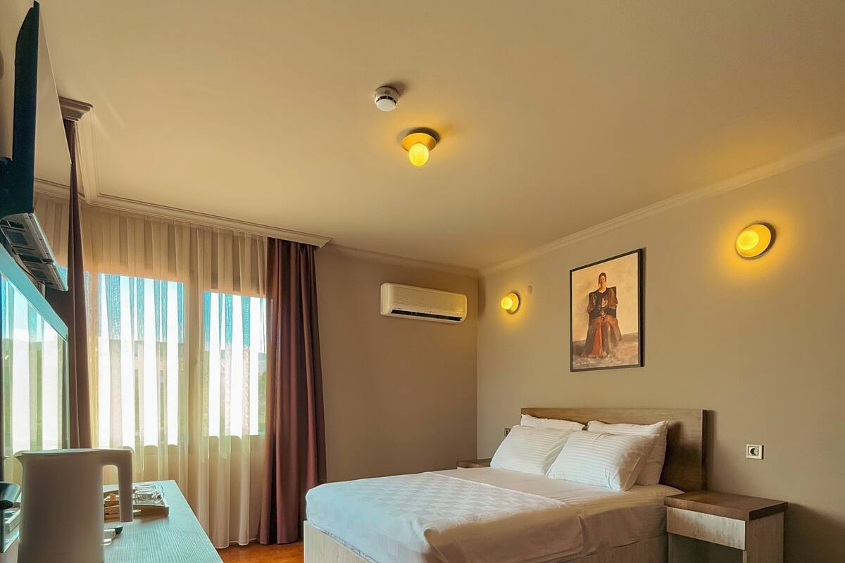 Ein Hotelzimmer mit Bett, Nachttischen, Klimaanlage, einem Gemälde an der Wand und einem Fenster mit Vorhängen.