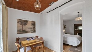 Una habitación con una mesa de comedor de madera, sillas, un cuadro, una cama y una lámpara colgante.