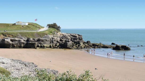 Een kuststrand met rotsachtige kliffen, een zandstrand en een grasheuvel met een wit gebouw en een vlag.