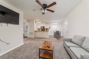 A living room with a ceiling fan, a gray sofa, a wooden coffee table, and a kitchen area in the background. - Budget 1BR Near Ft. Moore & Airport w. HDTV & WiFi (Columbus)
