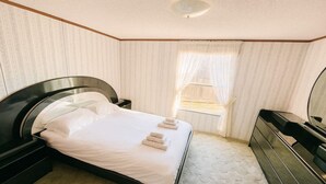 Un lit bien fait, avec du linge de lit et des serviettes blanches, une table de chevet, une commode et une fenêtre avec des rideaux.