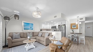A modern living room with a beige sofa, a wooden chair, a coffee table, and a dining area with a small table and chairs.