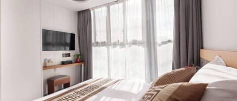 A modern hotel room with a large bed, a flat-screen TV mounted on the wall, a small desk with a chair, and a large window with curtains.