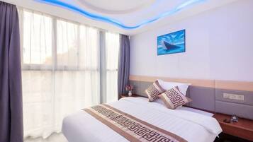 Quarto de hotel com cama grande, mesa de cabeceira, televisão na parede e vista através de cortinas finas.
