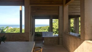 Una habitación con una mesa, sillas, un banco y vistas al océano y la vegetación.
