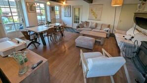 A spacious living room with a wooden floor, a sofa, a coffee table, and a fireplace. There is a dining area with a table and chairs, and a kitchen area with a sink and a countertop. - Charming house - Swimming pool - 11 beds - 15 travellers - Pets OK - Cluny (Azé)