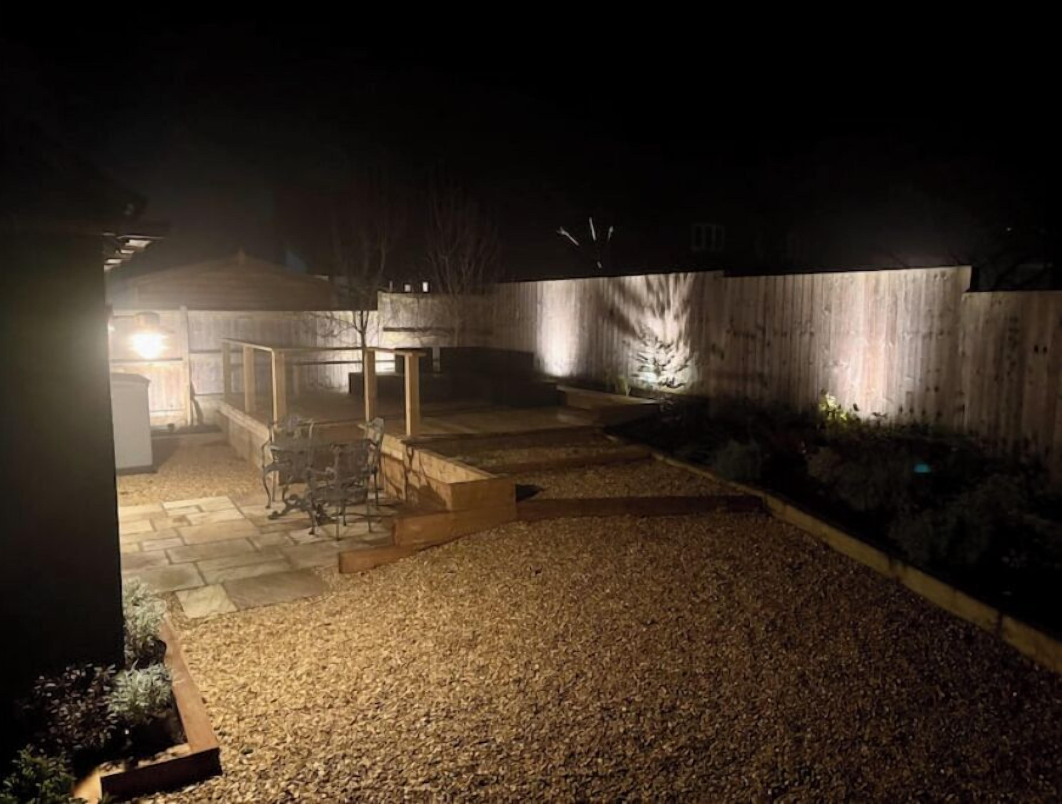 Een terras buiten, zwak verlicht, met een grindpad, houten banken en een kleine tafel.