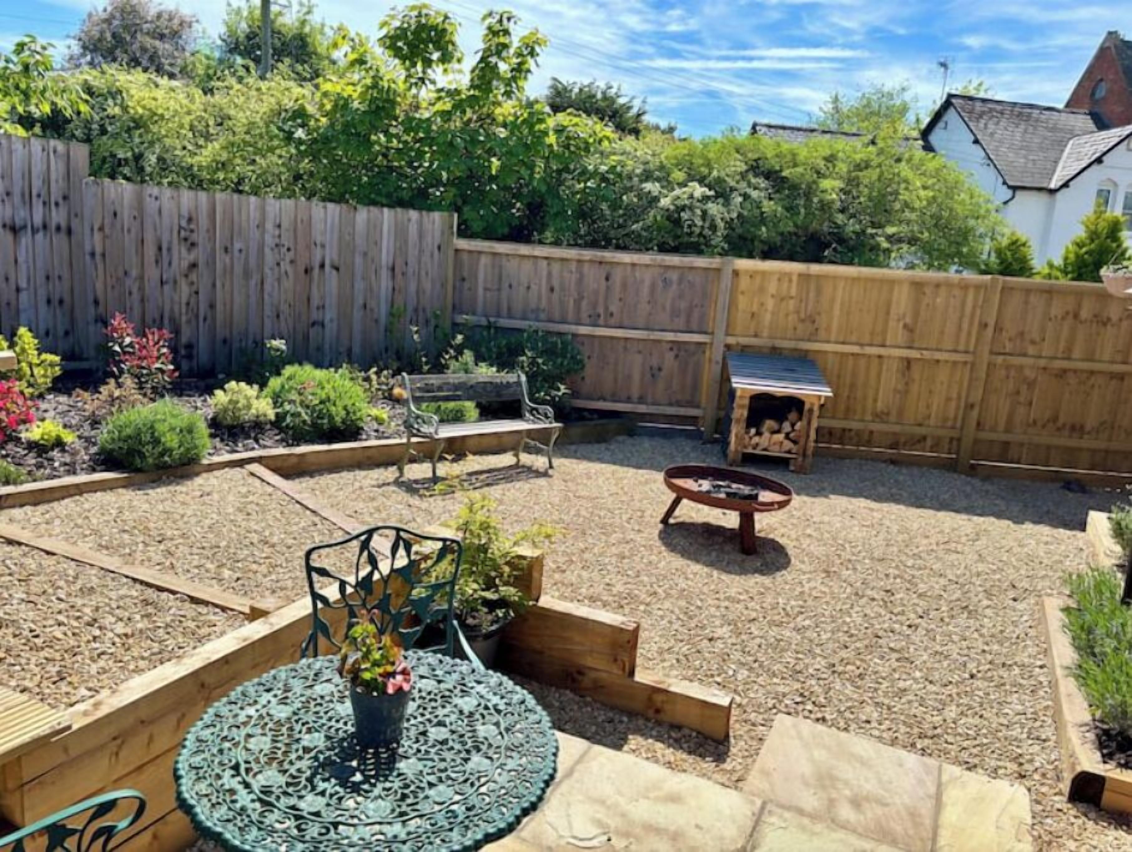 Un jardin à l’arrière de la maison, avec une terrasse en bois, un foyer, un banc et un petit espace aménagé en jardin, où diverses plantes sont cultivées.