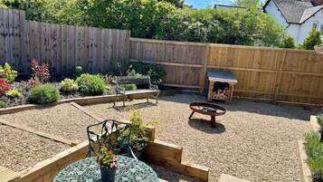 Un jardin à l’arrière de la maison, avec une terrasse en bois, un foyer, un banc et un petit espace aménagé en jardin, où diverses plantes sont cultivées.