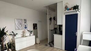 A modern living room with a white sofa, a coffee table, and a large mirror. There are several potted plants and a small kitchen area visible. - Studio Bassin de la Villette (Paris)