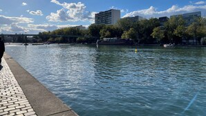 A riverside walkway with a bridge, trees, and buildings in the background. - Studio Bassin de la Villette (Paris)
