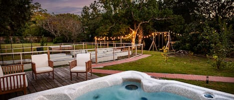 Un jacuzzi en una terraza de madera con sillas, rodeado por una valla y árboles, bajo un cielo crepuscular.