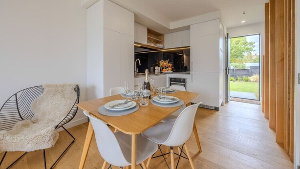 Un comedor moderno con una mesa de madera preparada para dos personas, sillas blancas y un sillón mullido. Se ve una cocina al fondo con vistas al exterior.