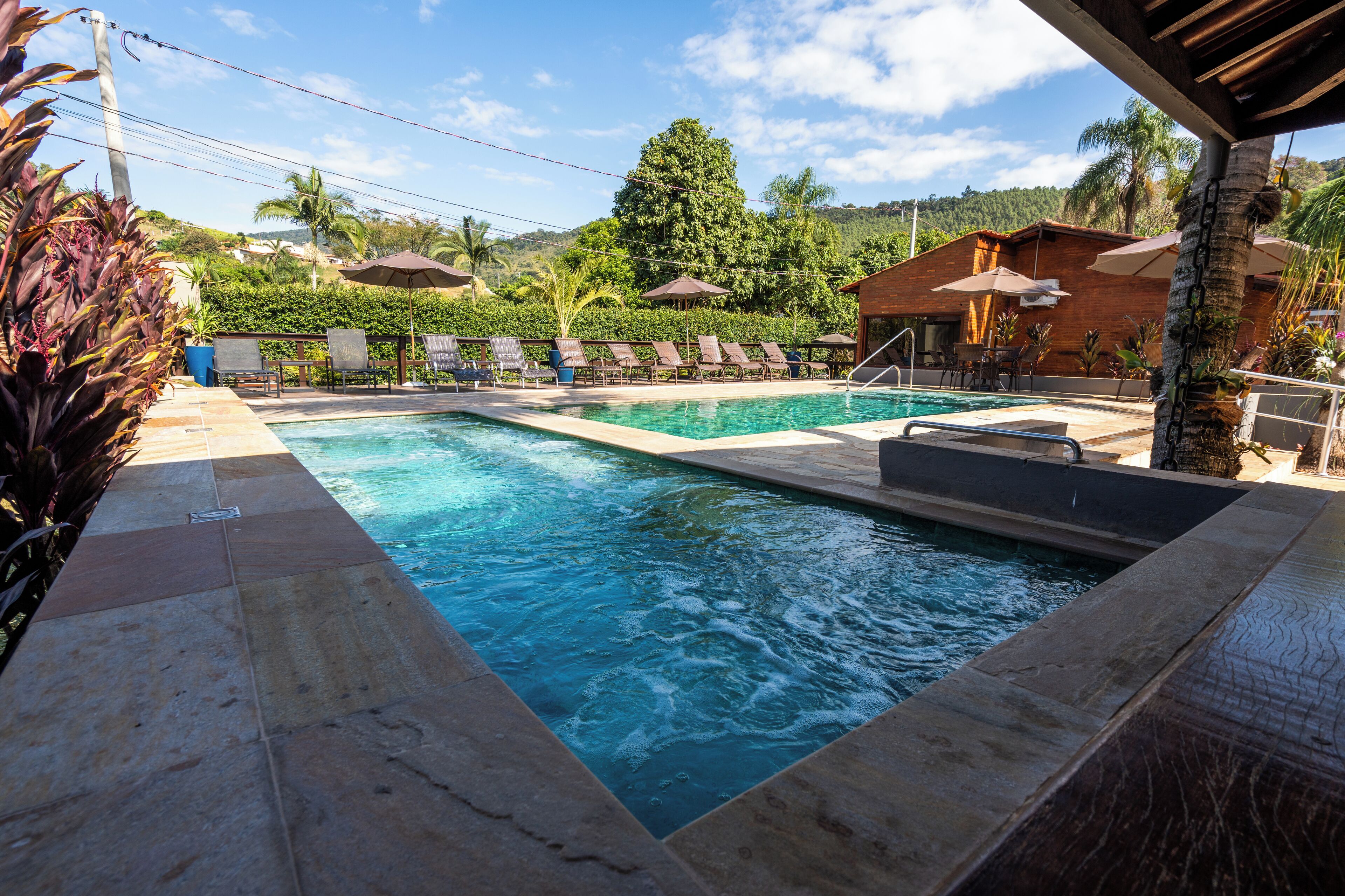 A swimming pool with lounge chairs and umbrellas, surrounded by greenery and a building.